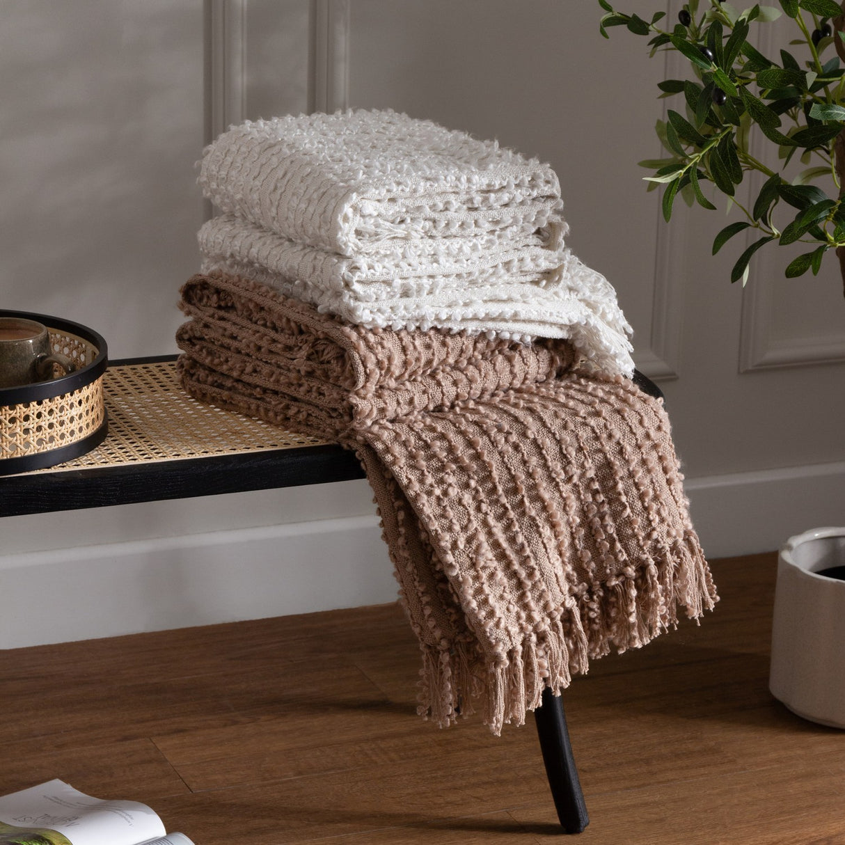 Arvo Plain Cotton Throw in natural, with textured boucle rows, folded on a black bench beside a fringed taupe throw in a living room setting.