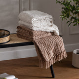 Arvo Plain Cotton Throw in natural, with textured boucle rows, folded on a black bench beside a fringed taupe throw in a living room setting.