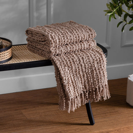 Arvo Plain Cotton Throw in Plaster, folded with fringed edges on a black cane bench, showing its ridged boucle-textured weave in a neutral living room setting.