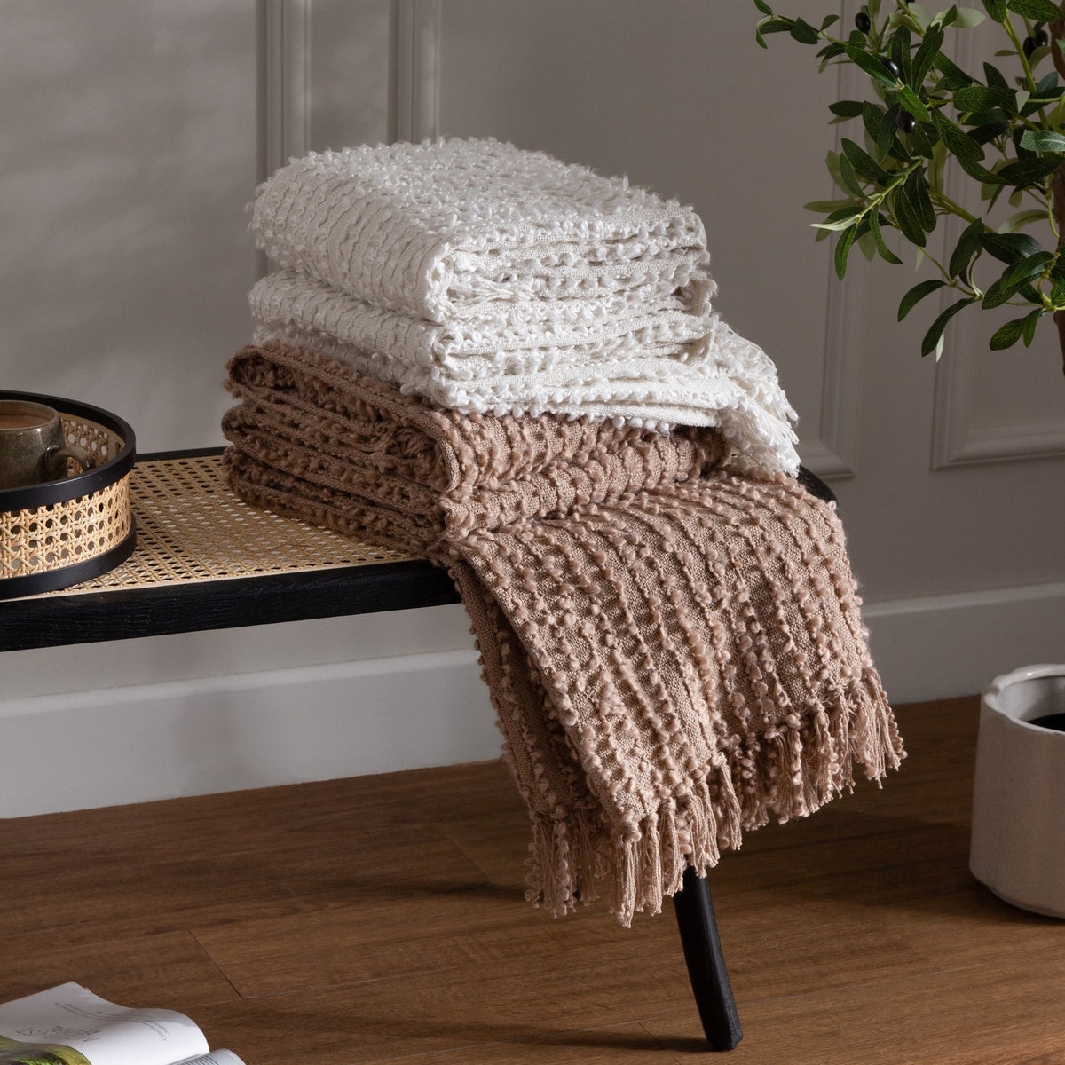 Folded Arvo Plain Cotton Throws in white and Plaster beige with textured boucle rows, stacked on a cane-topped bench with one throw draped and fringed.