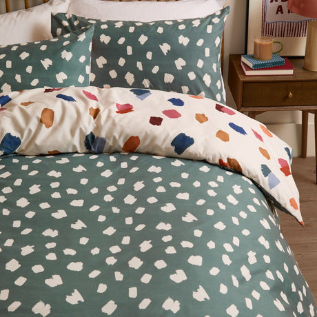 Astrid Abstract Duvet Cover Set on a bed, showing the reversible design with teal green cream-spot bedding and pillowcases in multicoloured brushstroke spots, in a bedroom setting.