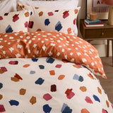 Astrid Abstract Duvet Cover Set on a bed, cream side with multicoloured brushstroke spots and reverse in orange with cream dots, shown with matching pillowcases in a bedroom setting.