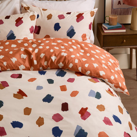 Astrid Abstract Duvet Cover Set on a bed, cream side with multicoloured brushstroke spots and reverse in orange with cream dots, shown with matching pillowcases in a bedroom setting.