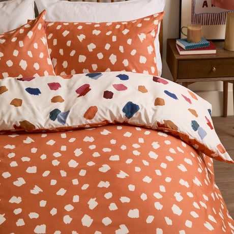 Astrid Abstract Duvet Cover Set in orange with cream brushstroke spots, reversing to a cream side with multicoloured marks, shown styled on a bed with matching pillowcases.