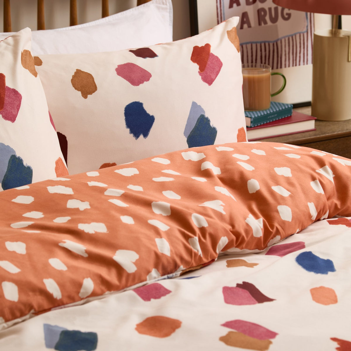 Astrid Abstract Duvet Cover Set on a bed, orange side with cream brushstroke spots shown alongside reverse cream pillowcase with multicoloured painted marks.