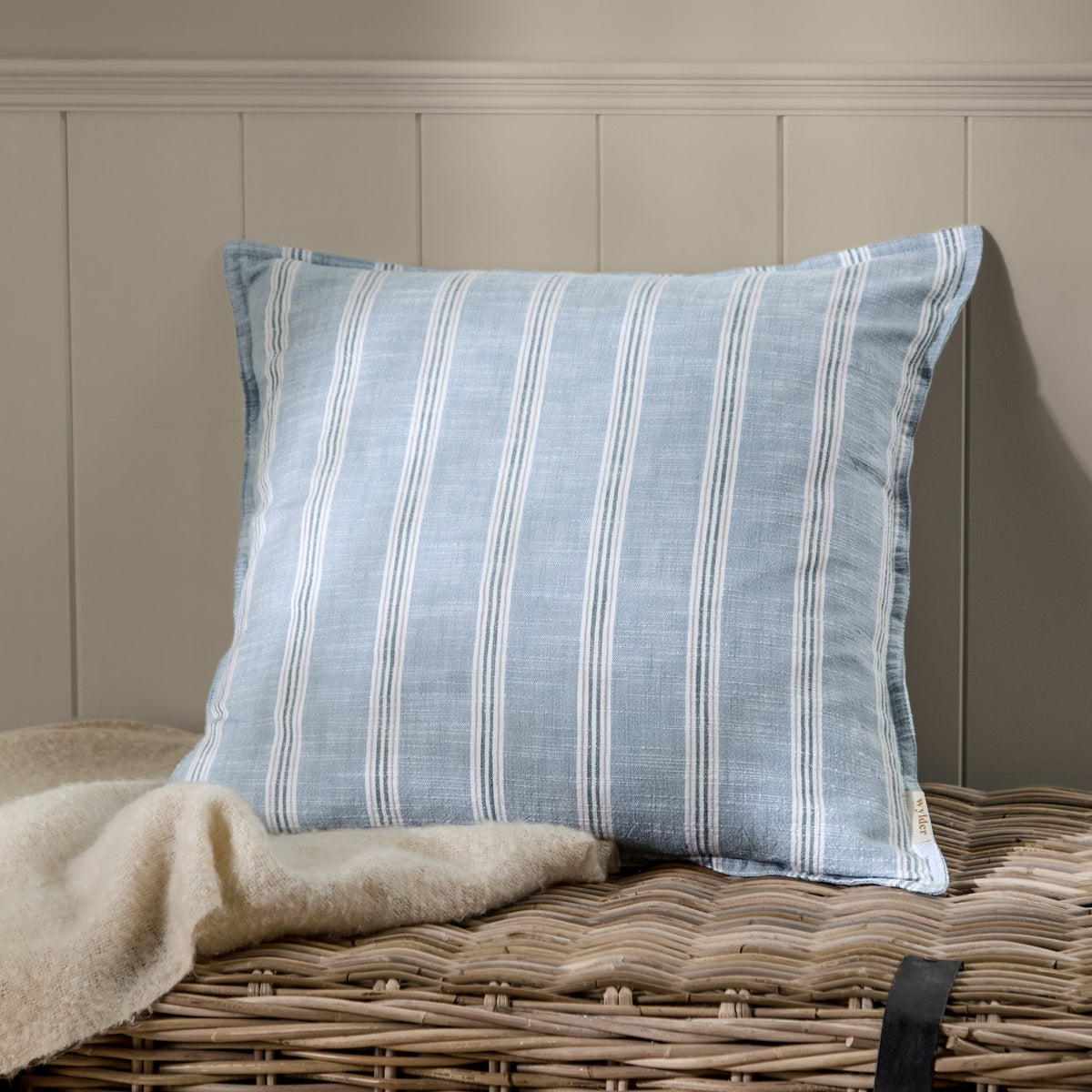 Abersoch Stripe Yarn-Dyed Cotton Cushion in Sail Blue with woven white stripes, shown on a wicker basket beside a cream throw against a panelled wall.