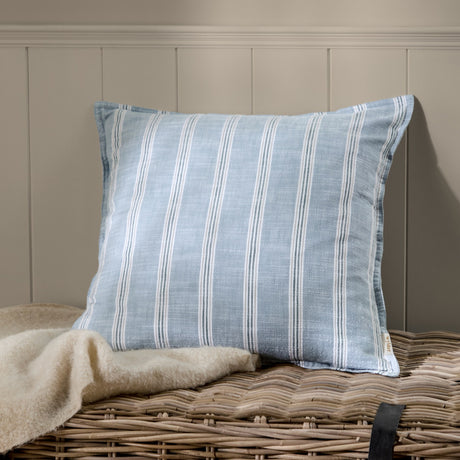 Abersoch Stripe Yarn-Dyed Cotton Cushion in Sail Blue with woven white stripes, shown on a wicker basket beside a cream throw against a panelled wall.