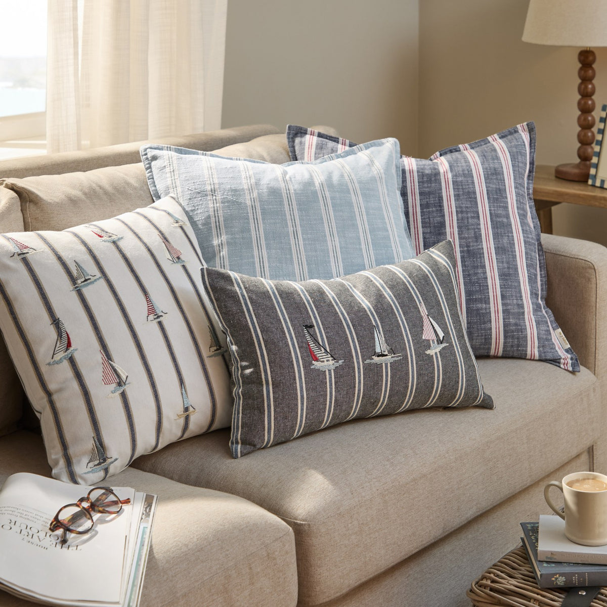 Abersoch Stripe Yarn-Dyed Cotton Cushion in Sail Blue, square with narrow woven stripes, shown among sailboat-print and grey striped cushions on a beige sofa.