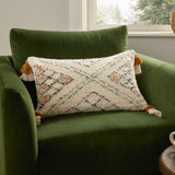 Atlas Tufted Cotton Cushion in Ochre, a rectangular bolster with cream, black and green diamond-chevron pattern and corner tassels, shown on a green velvet armchair.