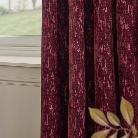 Aurelio Chenille Jacquard Fully Lined Eyelet Curtains in burgundy, shown in soft folds by a window, with a mottled woven texture and abstract pattern.