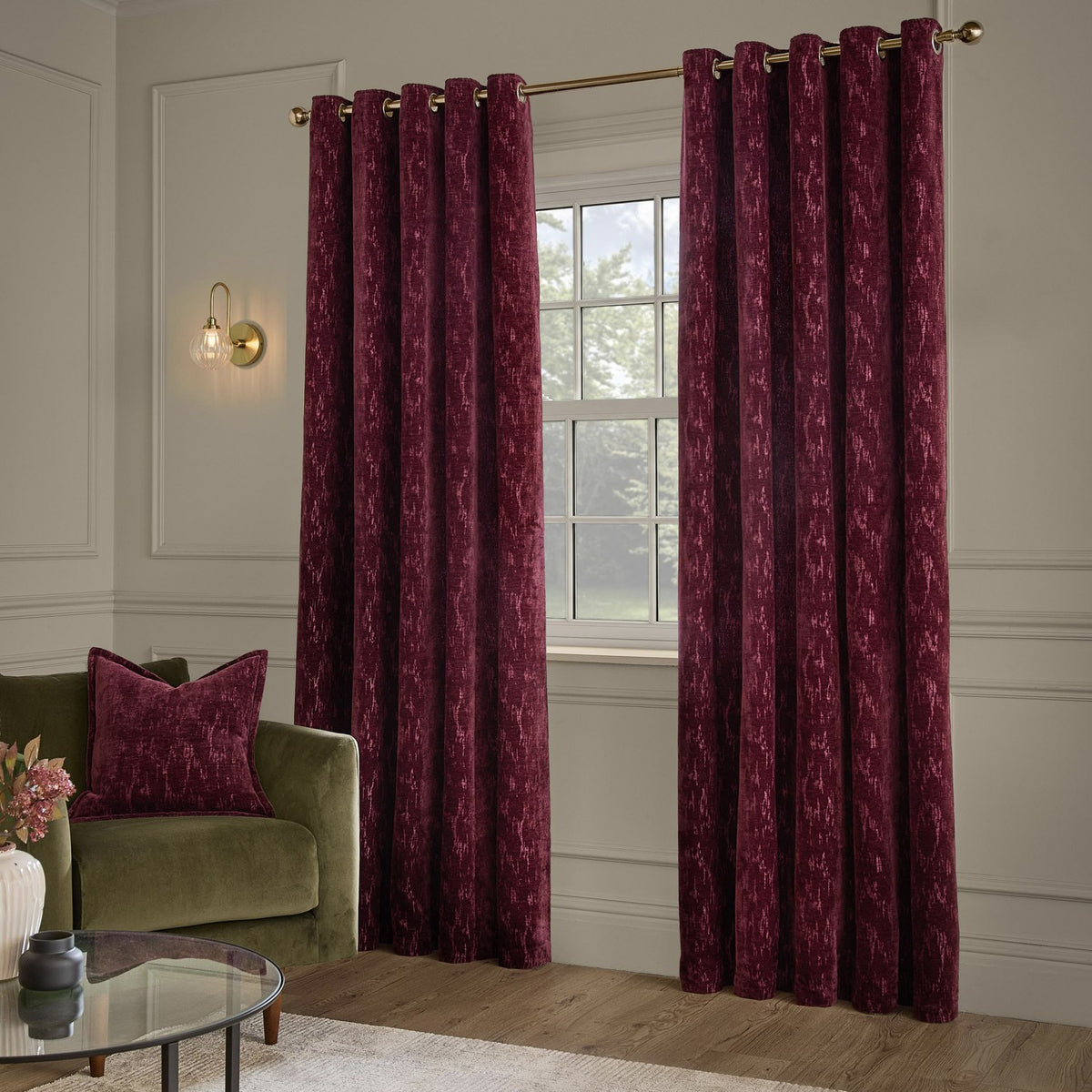 Aurelio Chenille Jacquard Fully Lined Eyelet Curtains in burgundy, shown hung on a brass eyelet pole either side of a window in a living room with a green sofa and burgundy cushion.