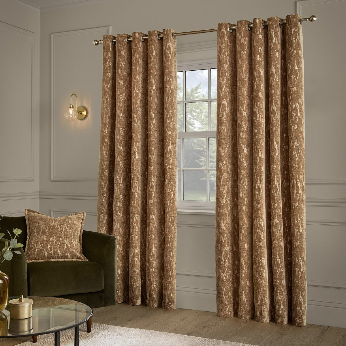 Aurelio Chenille Jacquard Fully Lined Eyelet Curtains in caramel with a mottled gold-toned pattern, shown on a metal pole in a living room with a green armchair.