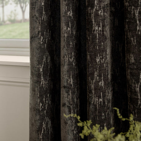Aurelio Chenille Jacquard Eyelet Curtains in charcoal, showing textured abstract jacquard weave with pale flecks, hung beside a window in a living room setting.