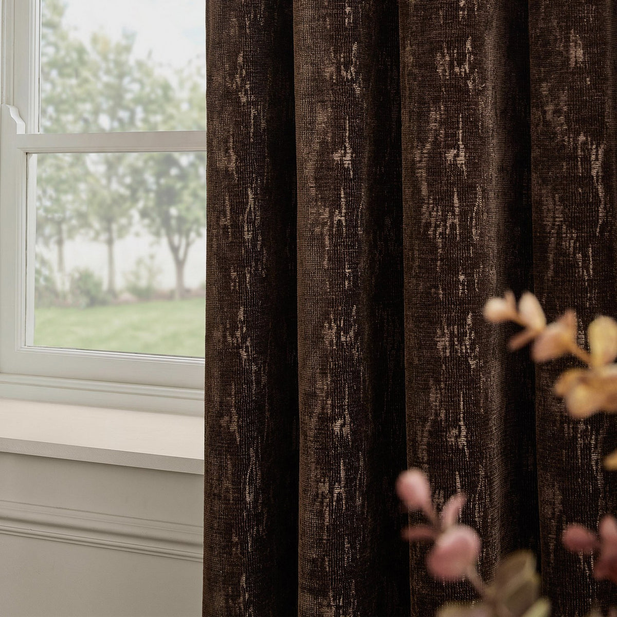 Aurelio Chenille Jacquard Eyelet Curtains in chocolate brown with subtle abstract pattern, shown hanging beside a white-framed window in a living room setting.