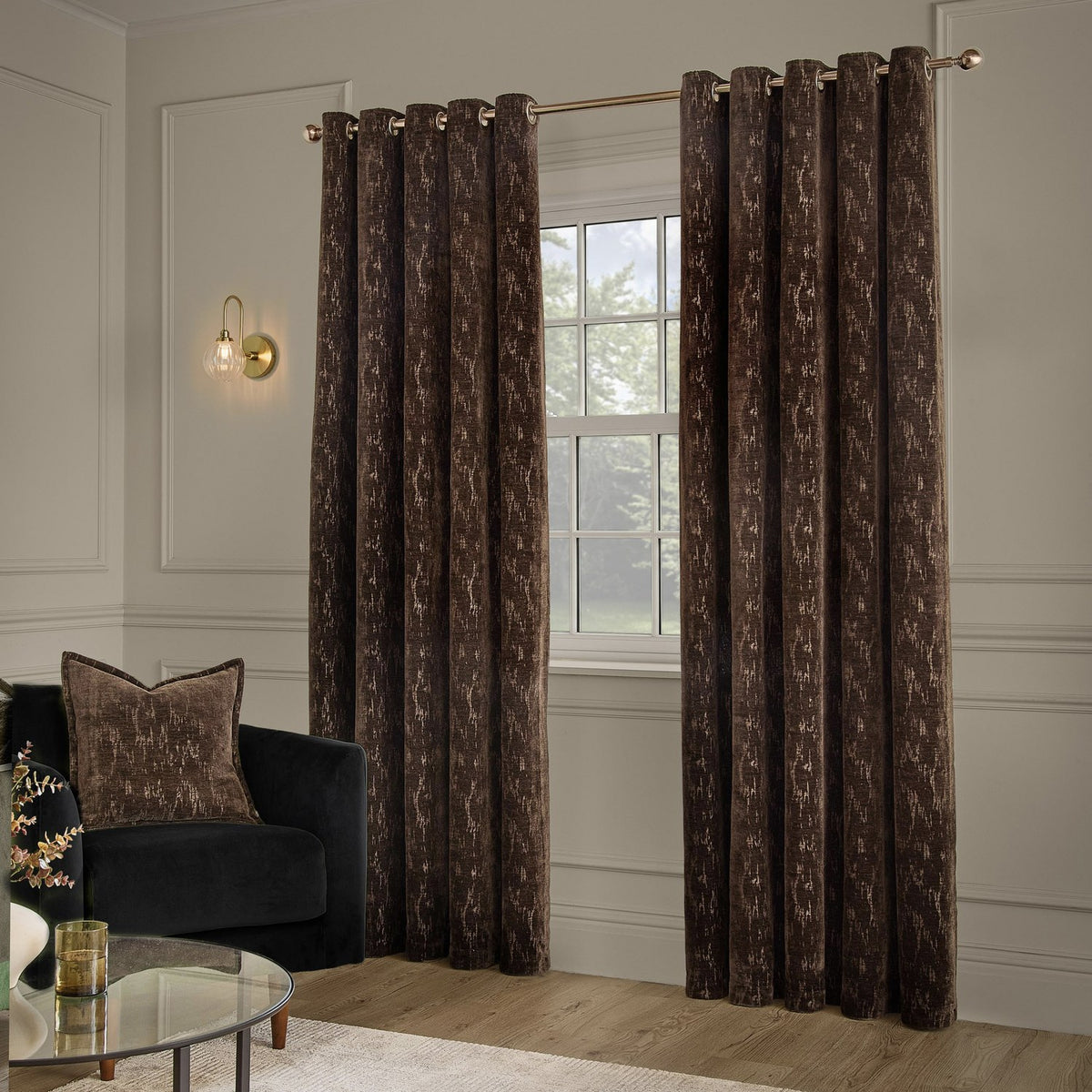 Aurelio Chenille Jacquard Fully Lined Eyelet Curtains in chocolate brown with subtle abstract pattern, shown hanging on a gold rod in a living room beside a window and dark armchair.