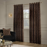 Aurelio Chenille Jacquard Fully Lined Eyelet Curtains in chocolate brown with subtle abstract pattern, shown hanging on a gold rod in a living room beside a window and dark armchair.
