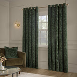 Aurelio Chenille Jacquard eyelet curtains in emerald green with a subtle abstract weave, shown hanging on a gold rod in a living room with a matching cushion on a brown sofa.