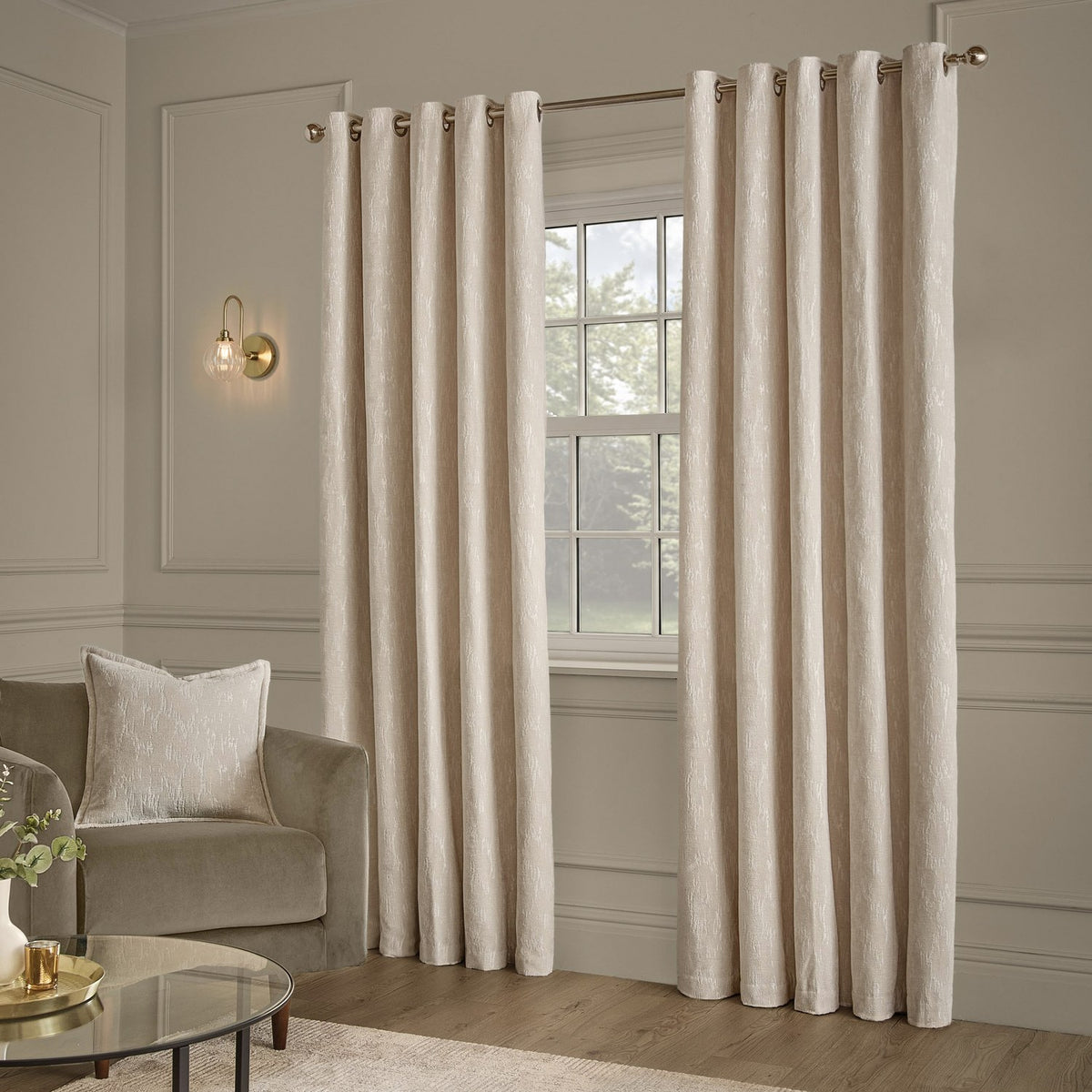 Aurelio ivory chenille jacquard eyelet curtains with brass rings, hung on a rod beside a window in a living room with panelled walls and a velvet armchair.