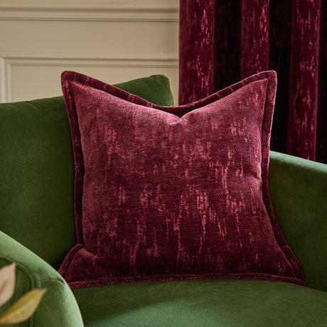 Aurelio Chenille Jacquard cushion in burgundy with a flanged edge and mottled woven texture, shown on a green velvet armchair against a burgundy curtain.