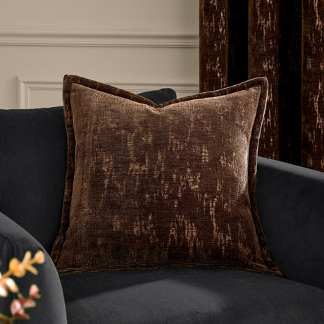 Aurelio Chenille Jacquard Polyester Cushion in chocolate brown with mottled abstract texture, styled on a dark velvet armchair against a neutral wall.