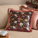 Autumn Garden Floral Velvet Cushion in Burnished Rose/Moss, square with pink fringed trim, on a beige sofa styled with pink cushions and books.