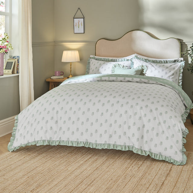 Ava Reversible Leaf Duvet Cover Set in White Sage on a bed with ruffled sage green trim, scattered leaf print, and matching pillowcases in a bedroom setting.