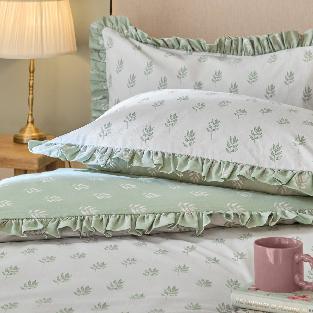 Ava Reversible Leaf Duvet Cover Set in White Sage, shown on a bed with ruffled pillow shams in white leaf print and sage green reverse, styled with a pink mug in a bedroom setting.