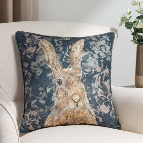 Avebury Animal Velvet Cushion in navy, square with a hare portrait in tan and amber tones amid pale florals, shown on a cream armchair.