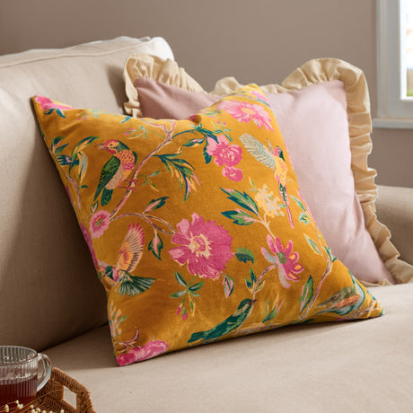 Aviary Velvet Cotton Cushion in gold with pink bird and floral print, shown on a beige sofa alongside pink and cream ruffled cushions.