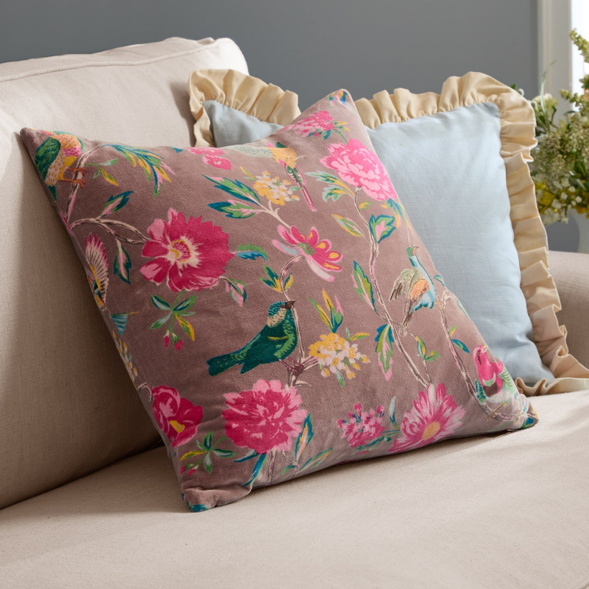Aviary Velvet Cotton Cushion in mink, a square cotton velvet cushion with a pink and green bird and floral print, shown on a beige sofa beside light blue and ruffled cushions.
