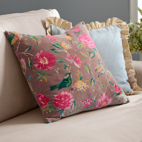 Aviary Velvet Cotton Cushion in mink, a square cotton velvet cushion with a pink and green bird and floral print, shown on a beige sofa beside light blue and ruffled cushions.