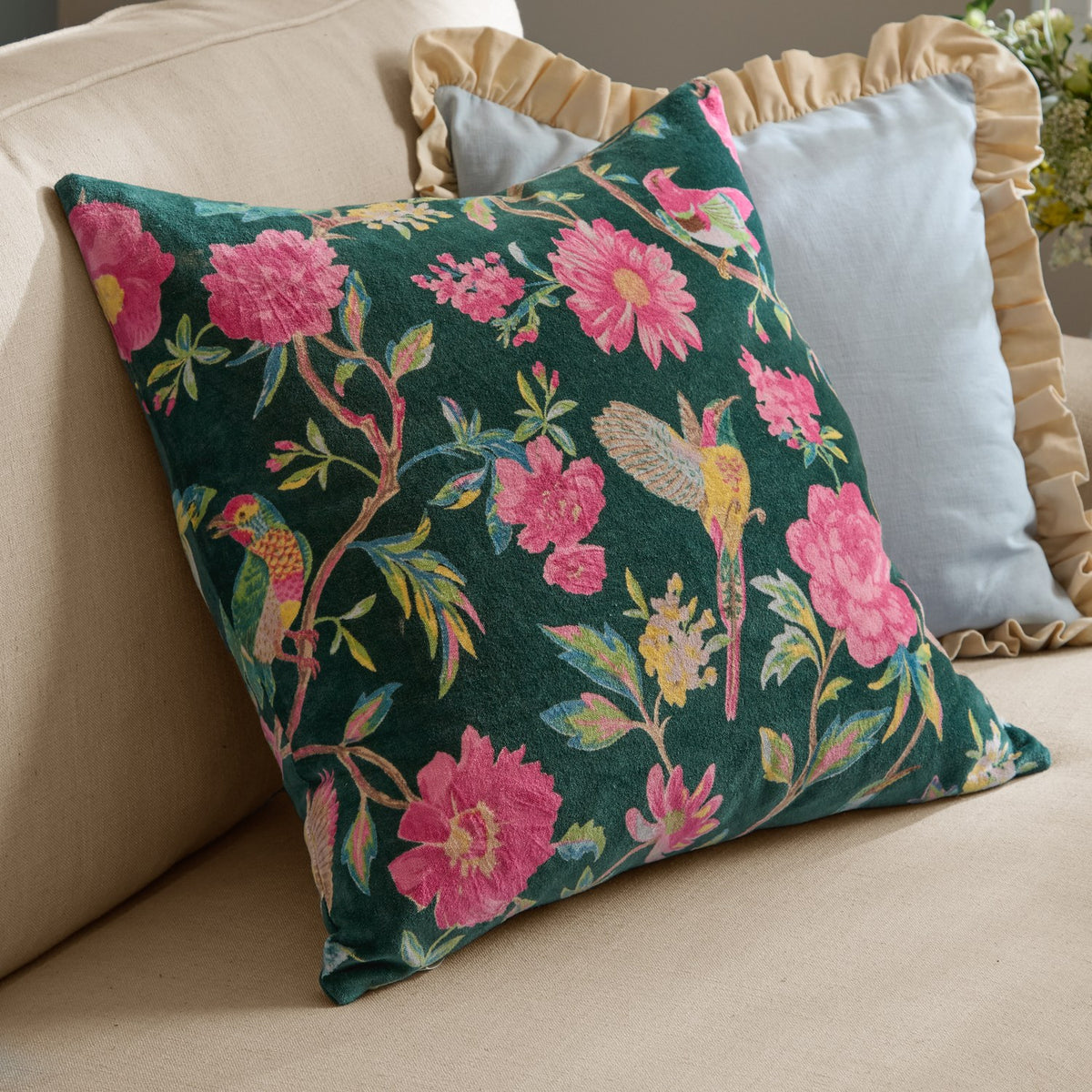 Aviary Velvet Cotton Cushion in teal green, square with pink floral and bird print, shown on a beige sofa beside light blue and gold ruffled cushions.