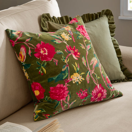 Aviary velvet cotton cushion in Woodgrove Green, square with bird and floral print in pink, yellow and teal, shown on a beige sofa beside a ruffled green cushion.