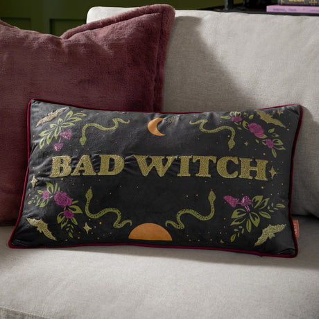 Rectangular black velvet cushion with red piping, "Bad Witch" in gold lettering, embroidered snakes, roses, bats and crescent moon, shown on a living room sofa.
