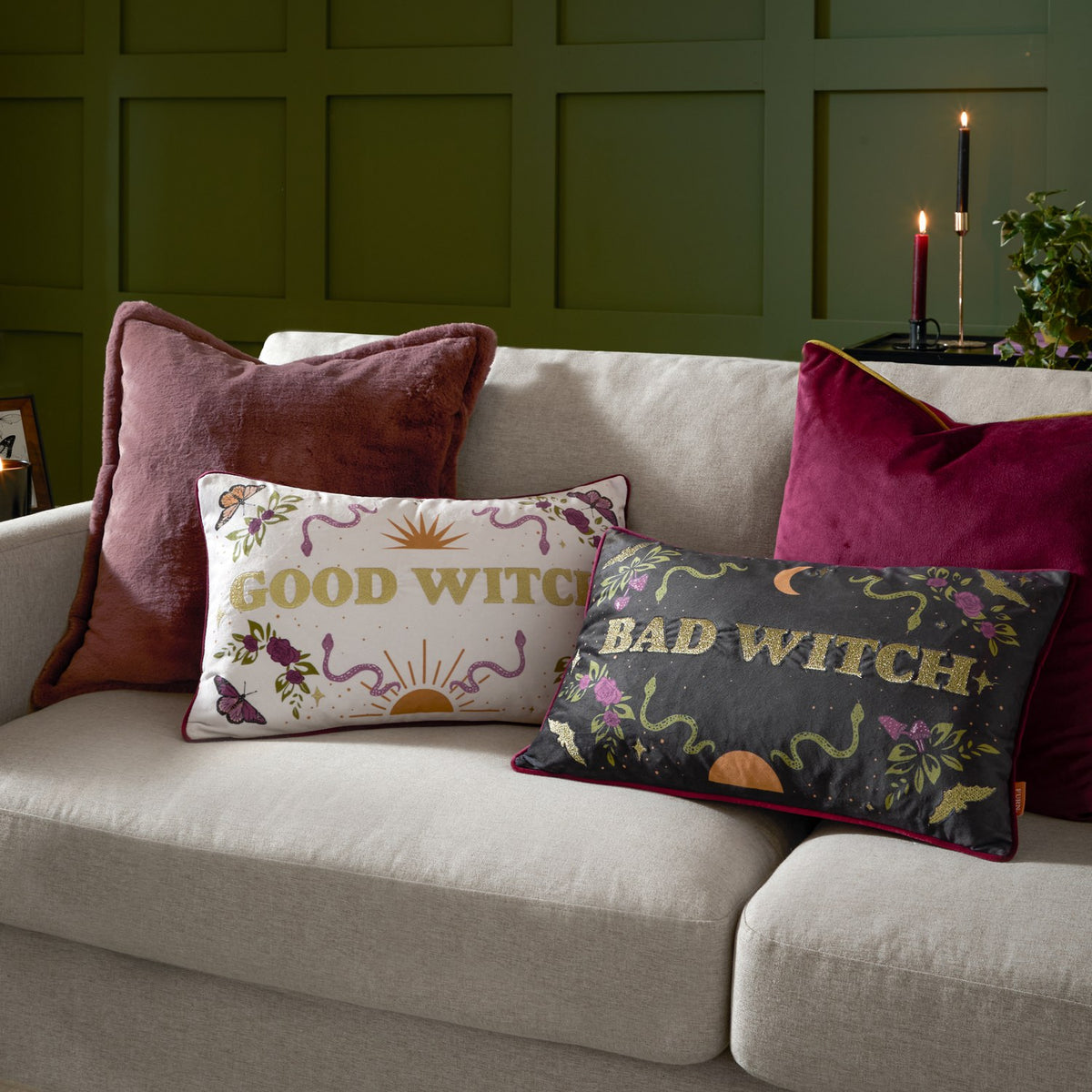Bad Witch Embroidered Velvet Cushion in charcoal with gold "Bad Witch" lettering, moon, sun, snake and floral motifs, styled on a beige sofa beside a Good Witch cushion.