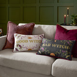 Bad Witch Embroidered Velvet Cushion in charcoal with gold "Bad Witch" lettering, moon, sun, snake and floral motifs, styled on a beige sofa beside a Good Witch cushion.