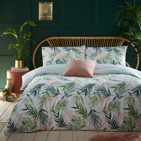 Bali Palm Reversible Botanical Duvet Cover Set in green on a bed with rattan headboard, white ground with palm leaf print, styled with a pink cushion in a green-panelled bedroom.