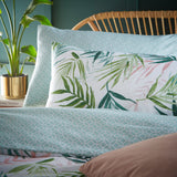 Bali Palm reversible duvet cover set styled on a bed, showing green palm leaf print pillowcases against a soft aqua geometric reverse, in a bedroom setting with rattan headboard and potted plant.