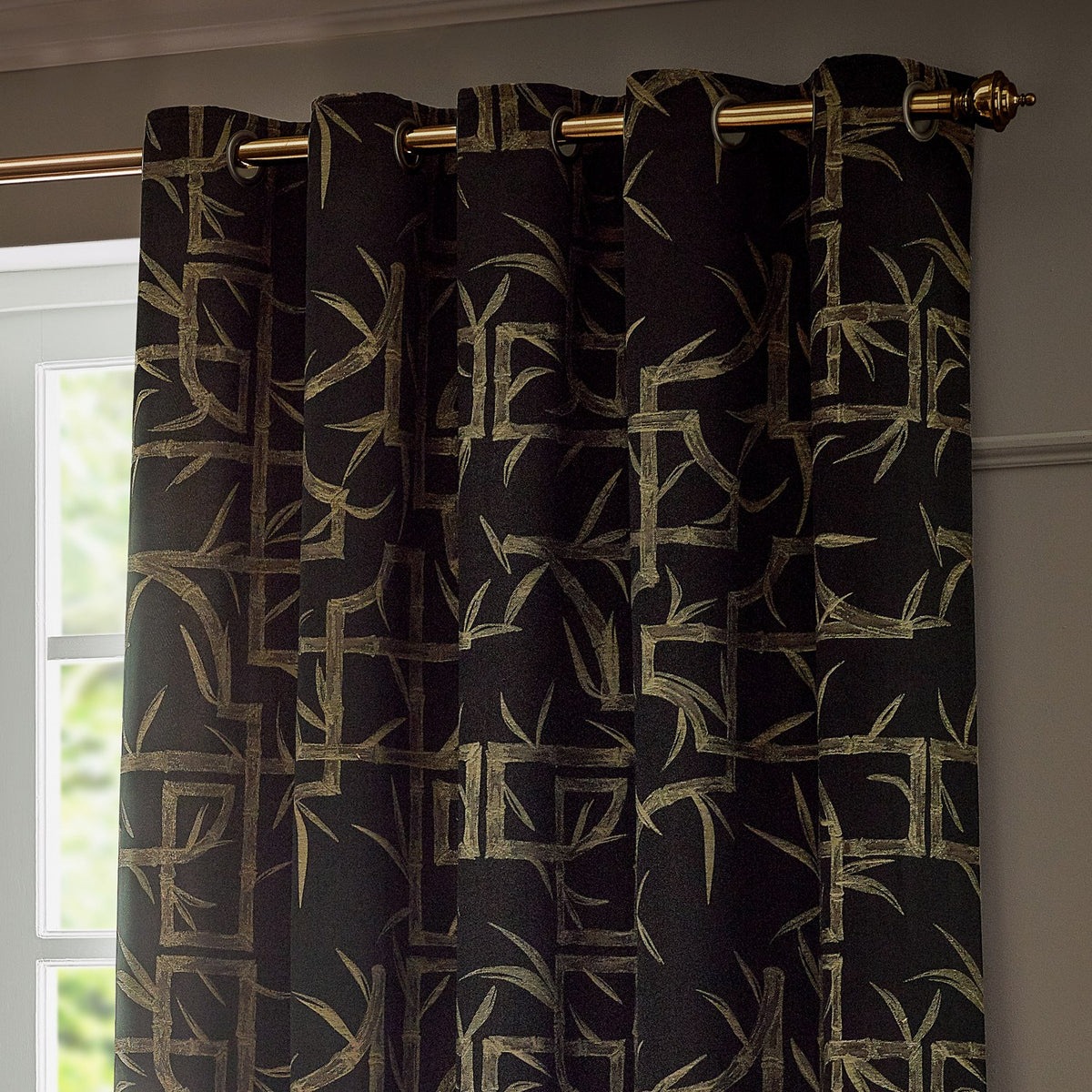 Bamboo Jacquard Lined Eyelet Curtains in black with pale gold bamboo cane and leaf pattern, hung on a brass pole with eyelet heading in front of a window.