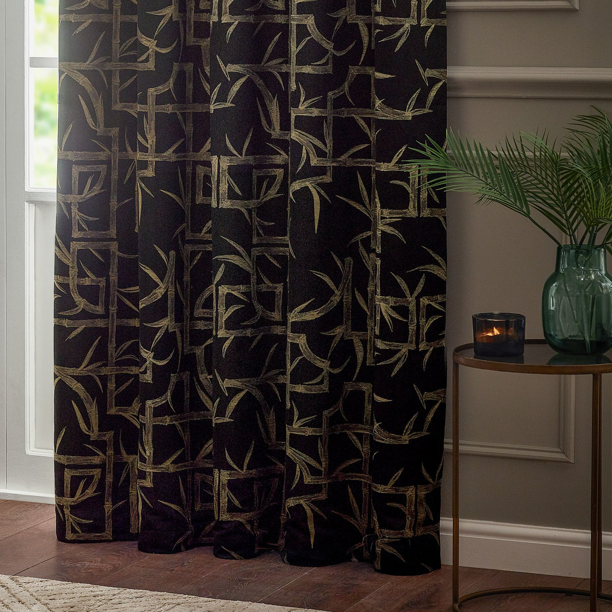 Bamboo Jacquard Lined Eyelet Curtain in black with pale gold bamboo cane and leaf pattern, shown hanging in a living room beside a green glass vase and side table.