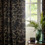 Bamboo Jacquard Lined Eyelet Curtains in black with pale gold-tan bamboo pattern, hung in eyelet folds beside a window overlooking greenery.