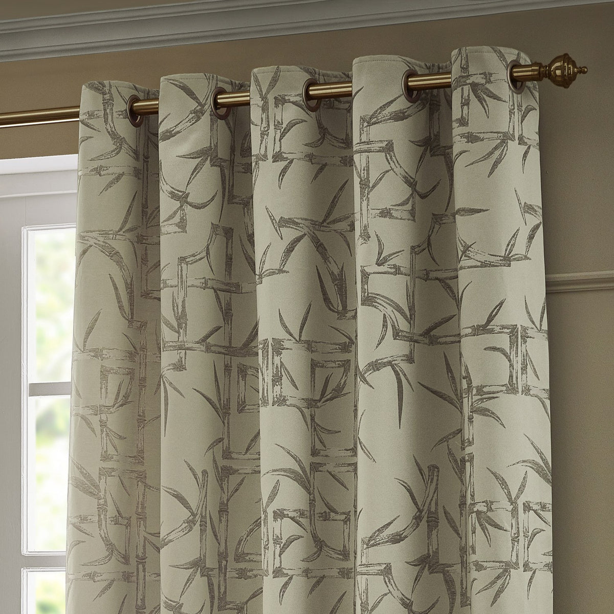 Champagne eyelet curtains with a jacquard bamboo cane and leaf pattern in soft grey, hung on a brass pole in a room setting.