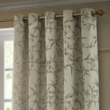 Champagne eyelet curtains with a jacquard bamboo cane and leaf pattern in soft grey, hung on a brass pole in a room setting.
