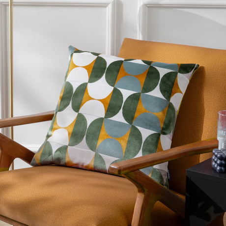 Bardot Geometric Velvet Cushion in gold, green, and blue-grey, a 50x50cm square cut-velvet design shown on an orange armchair.