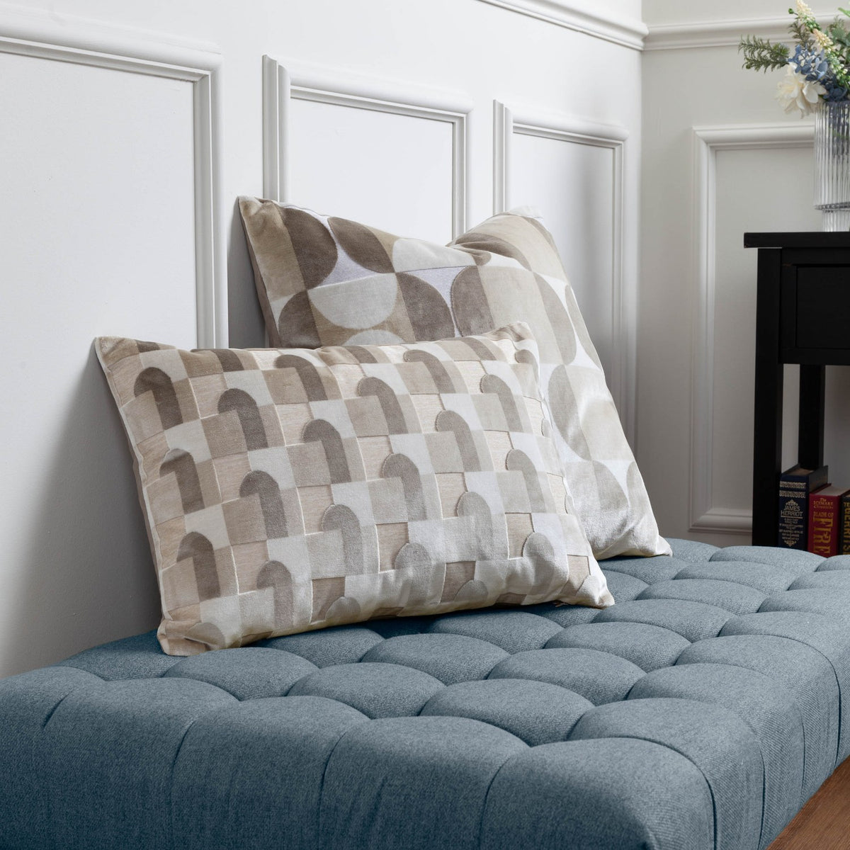Bardot Geometric Velvet Cushion in natural tones, shown with a coordinating patterned cushion on a blue tufted bench against white panelled walls.