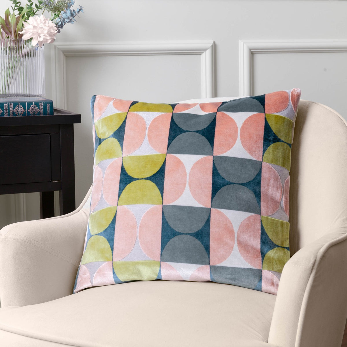 Bardot Geometric Velvet Cushion in pink and avocado green, cut-velvet half-circle pattern with slate blue, navy and cream, styled on a cream armchair beside a side table.