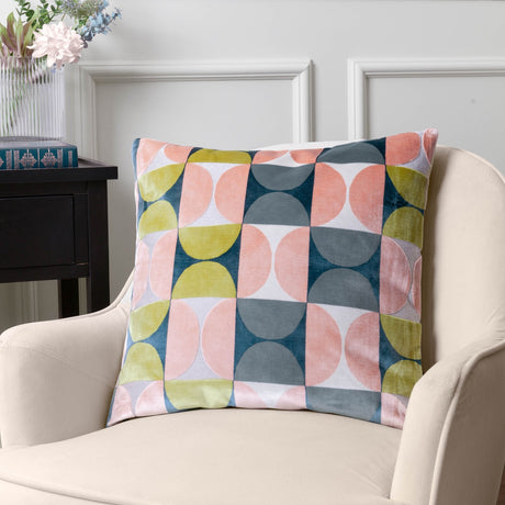 Bardot Geometric Velvet Cushion in pink and avocado green, cut-velvet half-circle pattern with slate blue, navy and cream, styled on a cream armchair beside a side table.