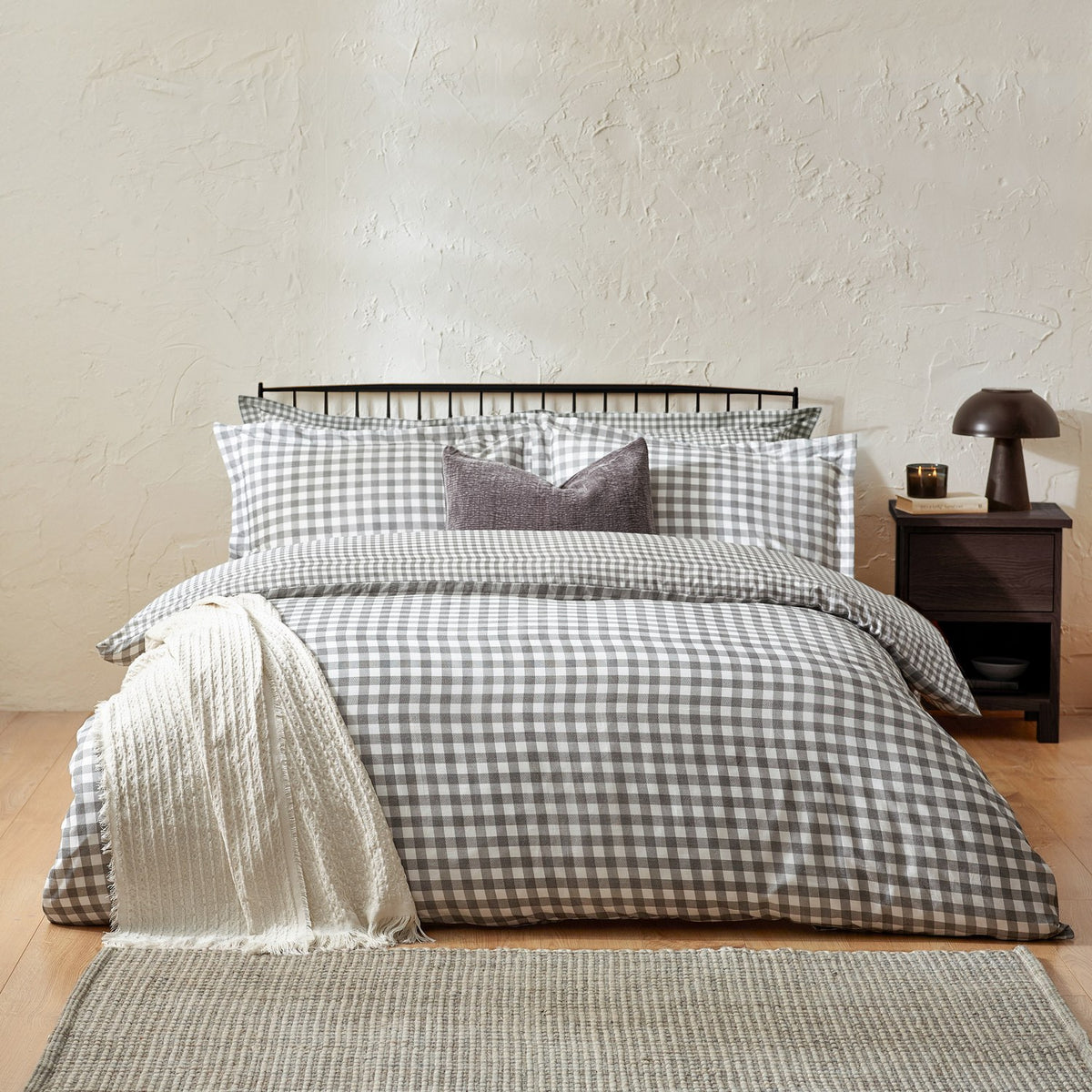 Barton Cotton Gingham Duvet Cover Set in grey and white check on a bed with matching pillowcases, a purple cushion, and a cream throw in a bedroom setting.