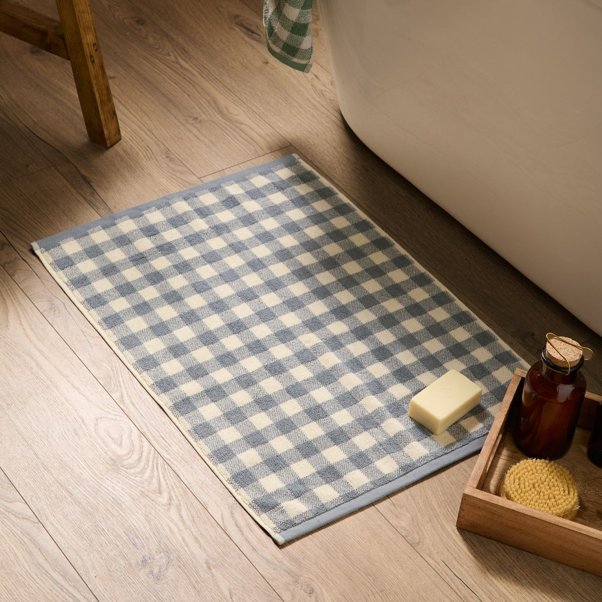 Barton Gingham Cotton Bath Mat in denim blue and cream check, shown on a wooden bathroom floor beside a freestanding bath, soap, and wooden tray.