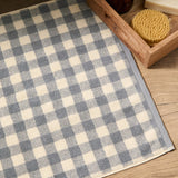 Barton Gingham Cotton Bath Mat in denim blue and cream check, shown on a wooden floor beside a bathroom tray with a soap bar and body brush.